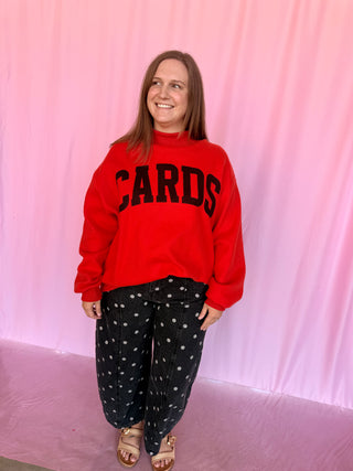A woman modeling a bright red sweatshirt with 'CARDS' printed in black collegiate letters across the chest, paired with black pants featuring white polka dots, standing against a pink draped backdrop.