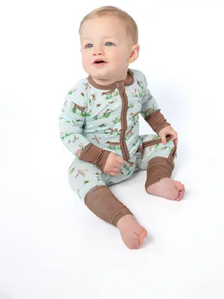 A baby sitting on a white studio background wearing a long-sleeve footless pajama romper with a light sage green forest pattern and brown trim.