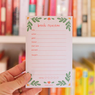 Hand holding a book review notepad with a floral design against a blurred bookshelf background