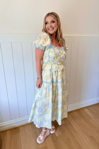 Woman wearing a floral dress standing in a room with wooden flooring and white walls.