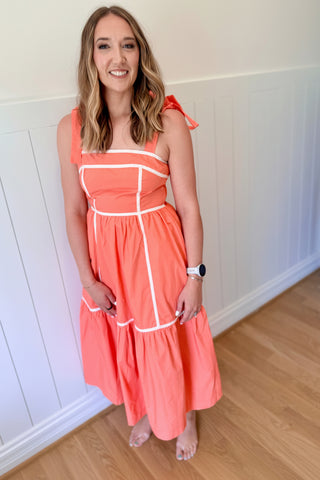 Woman wearing a coral dress with white accents in a room with wooden flooring and white walls.