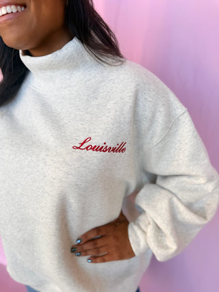 A close-up of a person wearing a light heather gray mock-neck sweatshirt with 'Louisville' embroidered in red script on the chest, set against a pink background.