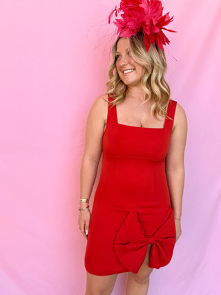 Woman wearing a red dress with a large bow and a pink headpiece against a pink background