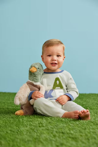 A toddler sitting on green artificial grass against a light blue backdrop, wearing a light gray long-sleeve pajama top with a large green and blue letter graphic and matching light gray pants. The child is holding a plush duck toy.