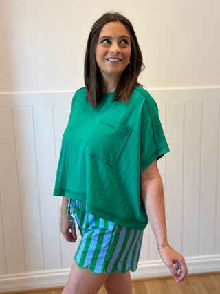 Woman wearing a green top and striped shorts against a white paneled wall.