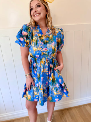Woman wearing a colorful dress with a floral pattern against a white paneled wall.