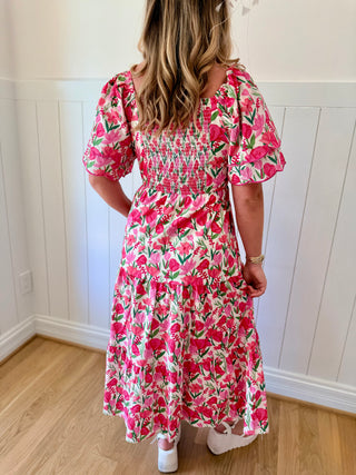 Woman wearing a pink floral dress standing in a room with wooden flooring and white walls.