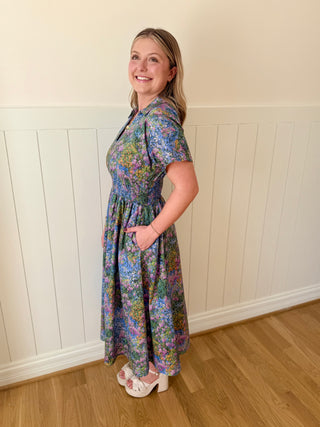 Woman wearing a floral dress standing against a white paneled wall.