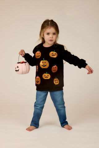 Child wearing a black sweater with Halloween pumpkin designs, holding a pink pumpkin-shaped basket.
