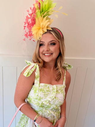Woman wearing a floral hat and green dress with a pink handbag indoors.