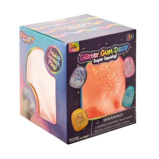 A boxed toy product labeled 'Glitter Gum Drop Super Squishy!' shown on a plain white background. The packaging is dark purple with colorful pastel graphics and a window revealing a peachy-orange glittery squishy material inside.