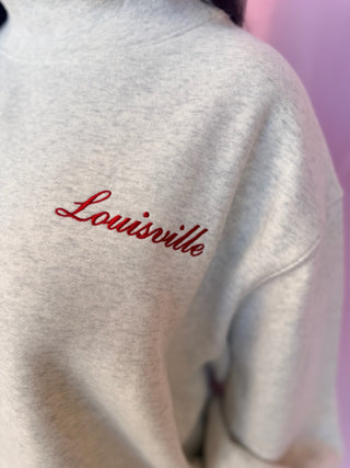 Close-up of a light heather gray sweatshirt with 'Louisville' embroidered in red cursive script on the chest.