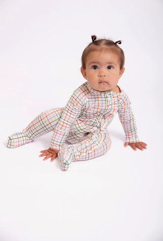 A baby girl sitting on a white studio background wearing a long-sleeve footed sleeper with a multicolor plaid pattern.
