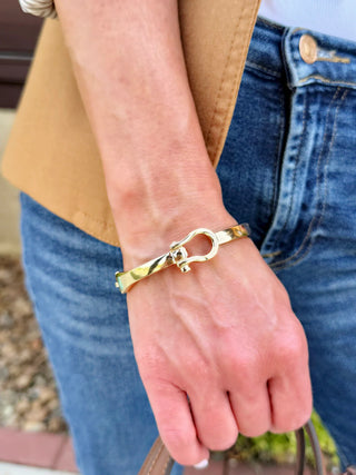 A close-up of a person's wrist wearing a polished gold-tone bangle bracelet with a distinctive open ring clasp, styled with blue jeans and a tan blazer.