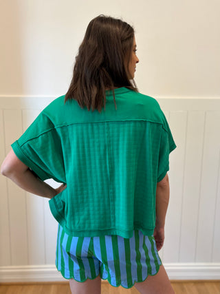Person wearing a green textured top and striped shorts against a white wall.