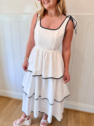 Woman wearing a white dress with black trim against a white paneled wall.