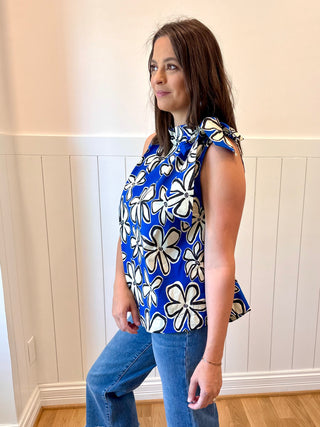 Woman wearing a blue floral top with white flowers, standing against a light-colored wall.