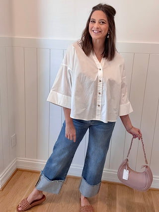 Woman wearing a white blouse and blue jeans, holding a pink handbag against a white paneled wall.