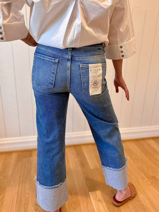 Person wearing blue jeans and a white shirt with a visible brand label, standing on a wooden floor.