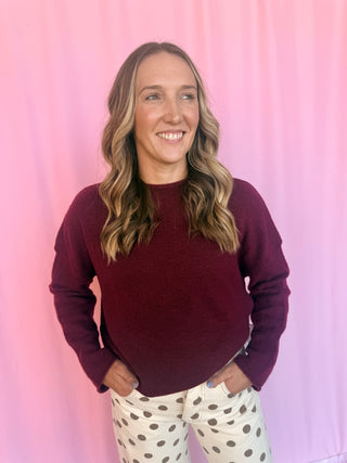 A woman with blonde-highlighted hair smiling, wearing a long-sleeve crewneck sweater in deep burgundy and cream pants with dark brown polka dots, standing against a pink curtain backdrop.