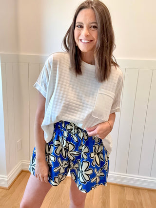 Woman wearing a white top and blue floral shorts in a room with wooden flooring and white walls.