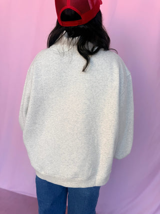 A person shown from behind wearing an oversized light heather gray sweatshirt, blue jeans, and a red trucker hat against a pink backdrop.