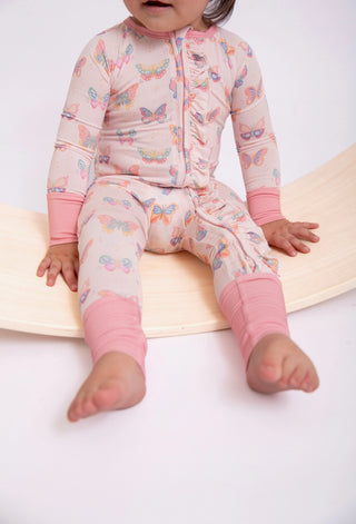 A baby wearing a light pink zip-up sleeper with a colorful butterfly print and solid pink cuffs and leg warmers, sitting on a curved wooden slide against a white background.