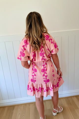 Woman wearing a pink floral dress with a large decorative headpiece against a white paneled wall.