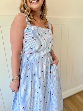 Woman wearing a blue checkered dress with cherry pattern against a white wall.