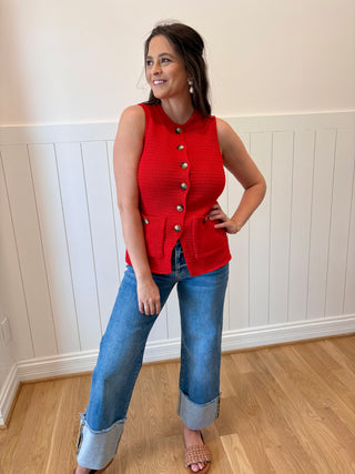 Woman wearing a red sleeveless top with buttons and blue jeans indoors.