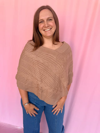 A smiling woman with brown hair stands against a pink backdrop wearing a chunky cable-knit poncho sweater in a warm taupe-brown color, paired with blue jeans.