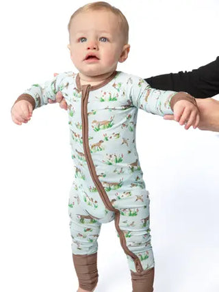 A baby wearing a long-sleeve footless zip-up sleeper with a brown and green woodland animal print on a pale blue background, held up by an adult's hands against a white studio background.