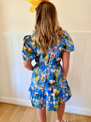 Woman wearing a blue floral dress standing against a white paneled wall.