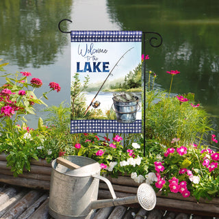 Decorative garden flag with fishing theme by a lake, surrounded by flowers and a watering can.