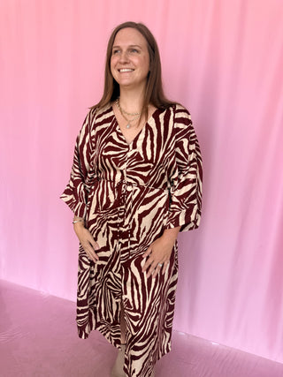 A woman modeling a zebra-print wrap dress with three-quarter sleeves, standing against a pink backdrop.