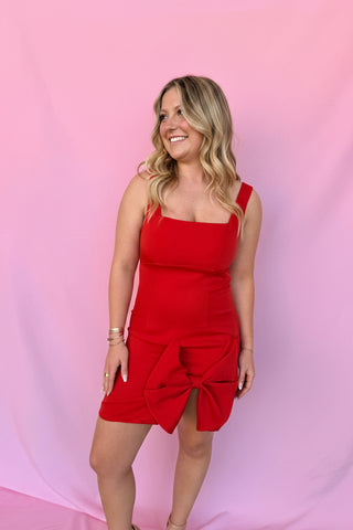 Woman wearing a red dress with a large bow and a matching red fascinator against a pink background