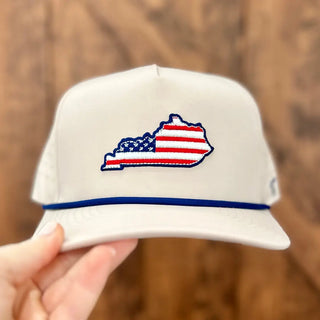 White cap with Kentucky-shaped American flag design held by a hand against a wooden background