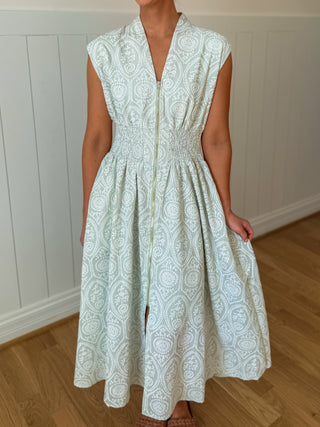 Woman wearing a light green sleeveless dress with a pattern against a white paneled wall.