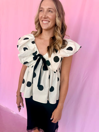 A woman wearing a white puff-sleeve blouse with black polka dots and a black bow at the neckline, paired with a black skirt, posing against a pink backdrop.