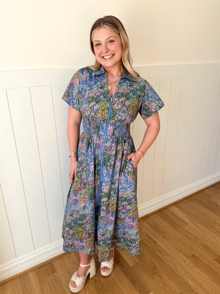 Woman wearing a floral dress standing against a white paneled wall.