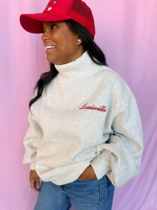 A woman wearing a light heather gray turtleneck sweatshirt with 'Louisville' embroidered in red on the chest, paired with a red trucker hat and blue jeans, posing against a pink backdrop.