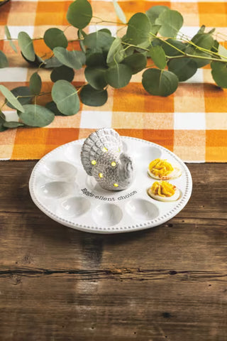 A white ceramic egg-cellent choice plate with molded egg slots, a gray snail-shaped egg holder in the center, and two hard-boiled eggs topped with yellow mustard, set on a rustic wooden table with an orange-and-white checkered cloth and eucalyptus leaves in the background.