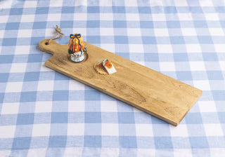 A rectangular wooden serving board with a rounded handle and a small hanging loop, placed on a blue-and-white checkered tablecloth. The board holds a small decorative orange-and-black ornament and a small white-and-orange item.