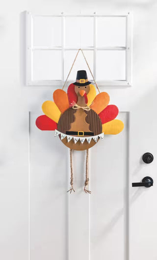 A handmade wooden Thanksgiving turkey hanging decoration on a white paneled door. The turkey features a brown striped body, a black Pilgrim hat, a red and yellow wattle, and multicolored feather shapes in red, orange, and yellow.