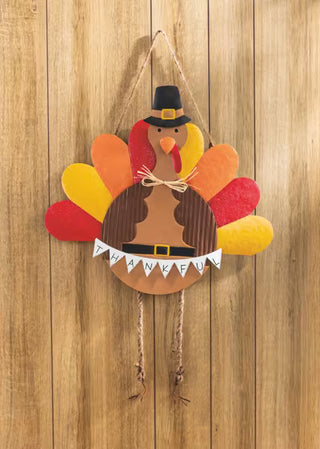 A handmade Thanksgiving turkey wall hanging decoration featuring a brown cardboard body, colorful felt feathers in red, orange, and yellow, a black pilgrim hat, and a 'THANKFUL' banner, hanging against a wooden plank wall.
