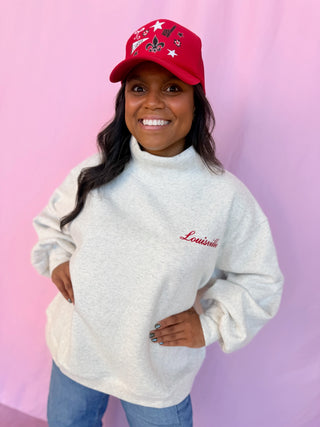 A smiling woman wearing a light heather gray turtleneck sweatshirt with 'Louisville' embroidered in red on the chest, paired with a red baseball cap and blue jeans, standing against a pink backdrop.