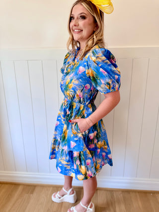 Woman wearing a blue floral dress standing against a white paneled wall.