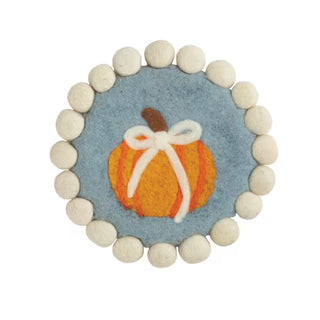 A round felt pin or brooch featuring a blue center with an orange pumpkin and white bow, surrounded by a border of cream pom-poms on a white background.