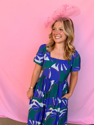 Woman wearing a blue and green patterned dress with a pink hat against a pink background
