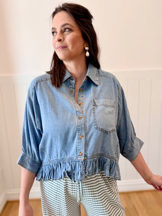 Woman wearing a denim shirt with ruffled hem in a room with a white wall.